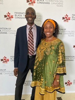 Clovis is wearing a blue blazer with a light dress shirt and a striped tie. Sherron is wearing a green and gold outfit with a bright orange hairband. They’re standing side-by-side while smiling at the camera.